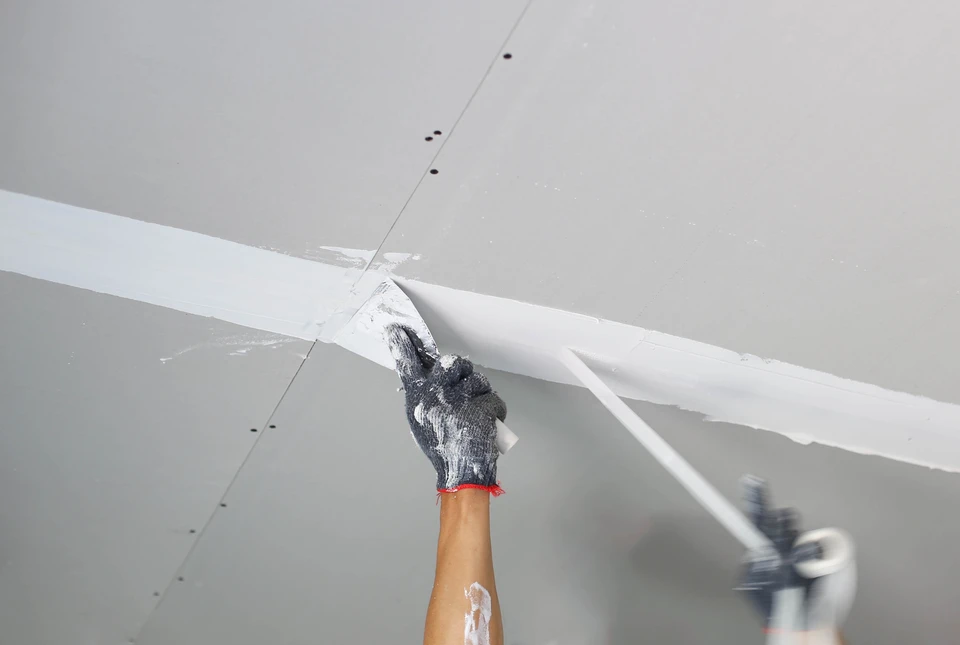 Une personne applique de l'enduit ou du mastic à l'aide d'un couteau à enduire sur une jonction entre plaques de plâtre au plafond