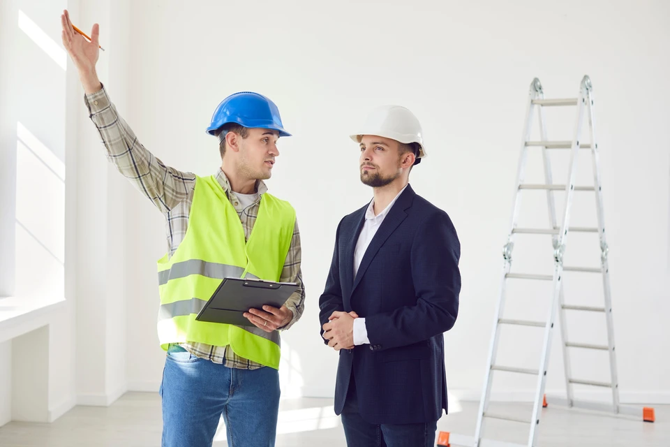 Contremaître discutant avec le jeune projet de conception d'architecte ou de réparation d'un nouvel appartement debout dans une pièce blanche vide sur le chantier de construction