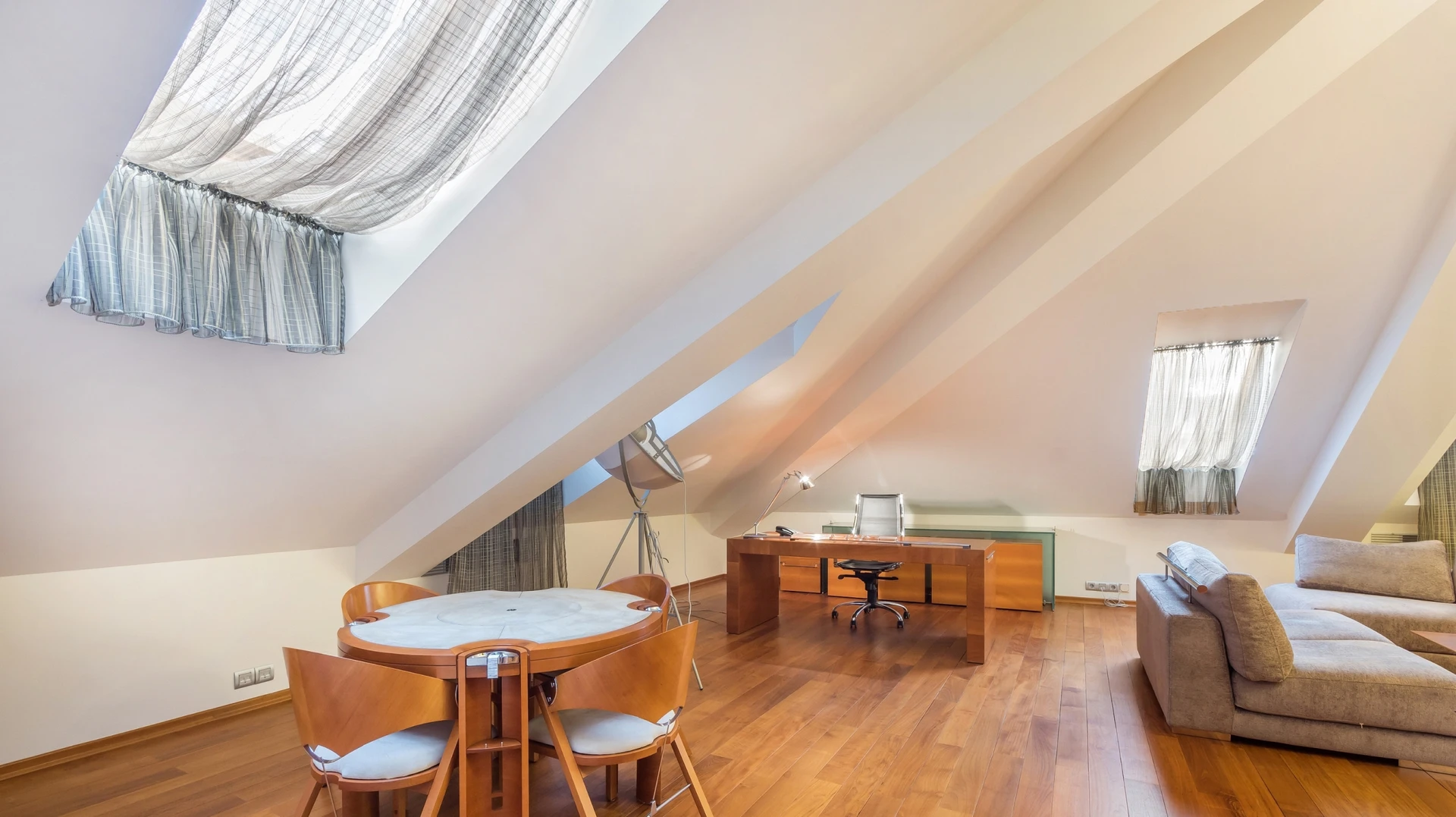 Chambre moderne sous combles avec poutres apparentes blanches, parquet en bois foncé, lit bas contemporain avec coussins gris, fauteuil en rotin blanc, console blanche avec miroir ovale à cadre noir, lampe de table dorée, velux apportant lumière naturelle